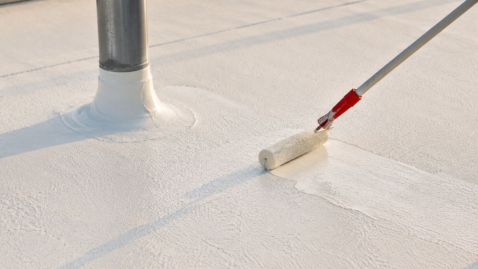 A paint roller applying a white protective coating to a flat roof around a metal pipe.